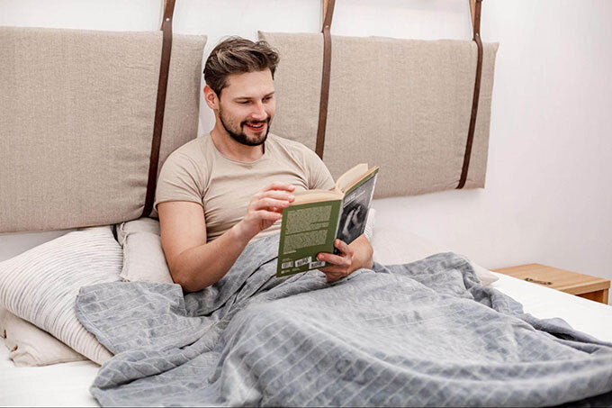 Mann liest entspannt im Bett unter einer grauen Gewichtsdecke